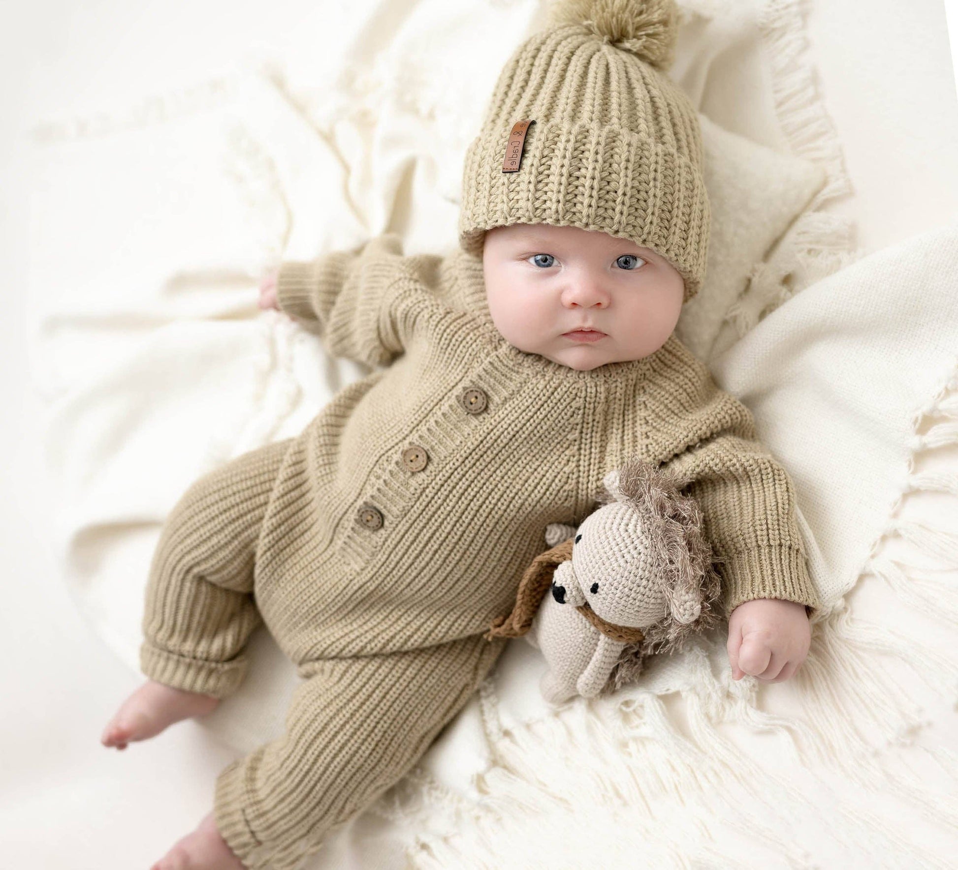 Baby in a beige knitted outfit and hat lying on a white blanket