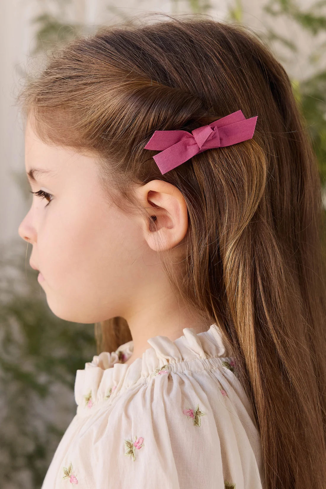 A young girl with long brown hair is shown in profile wearing a dusty pink fabric hair bow clip in her hair, paired with a cream-colored embroidered blouse.