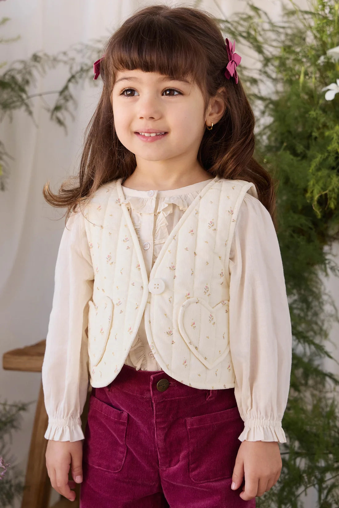A young girl with brown hair and pink bows is wearing a cream-colored floral embroidered vest over a cream blouse, paired with magenta corduroy pants, standing in front of a soft-focus indoor background with greenery.