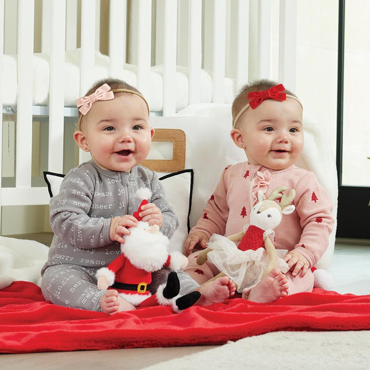 Two babies sitting on a bed with Christmas-themed stuffed toys and headbands.