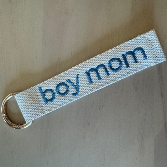 A white woven fabric keychain strap with blue embroidered text reading 'boy mom' and a gold metal key ring, placed on a light wood surface.