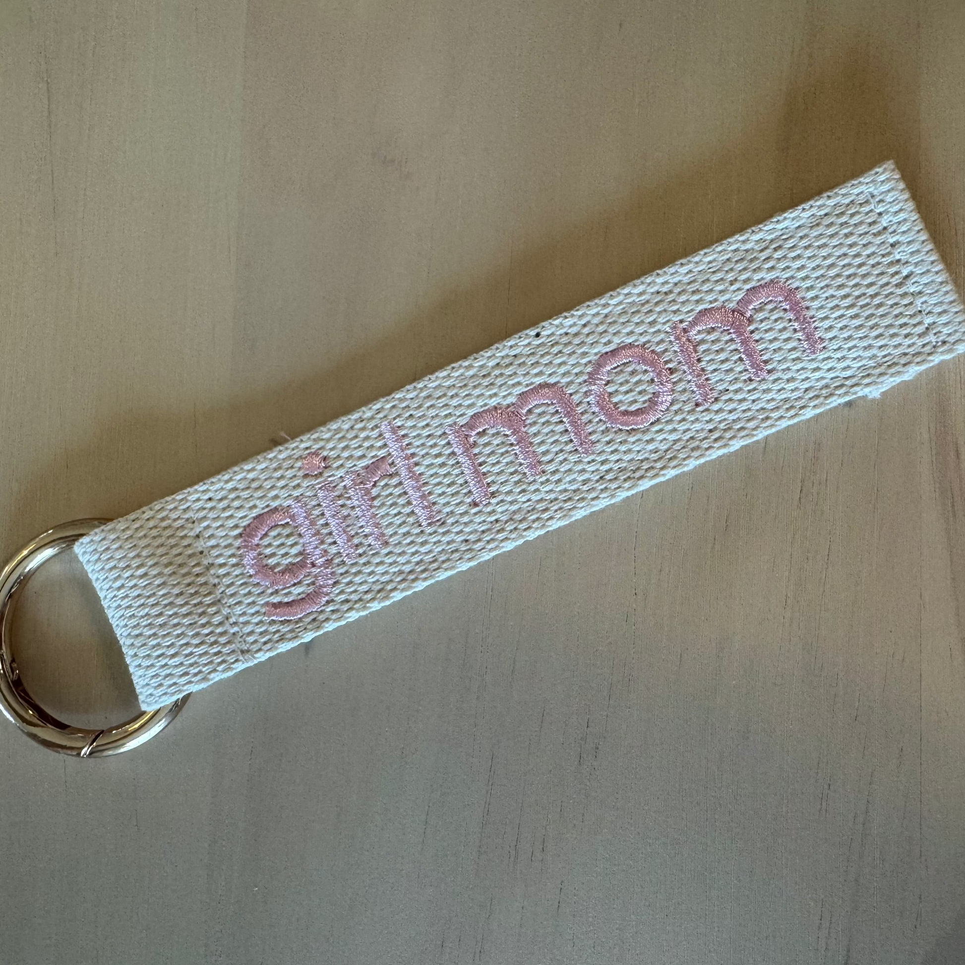 A white woven fabric keychain strap with pink embroidered text reading 'girl mom' and a gold metal key ring, placed on a light wood surface.