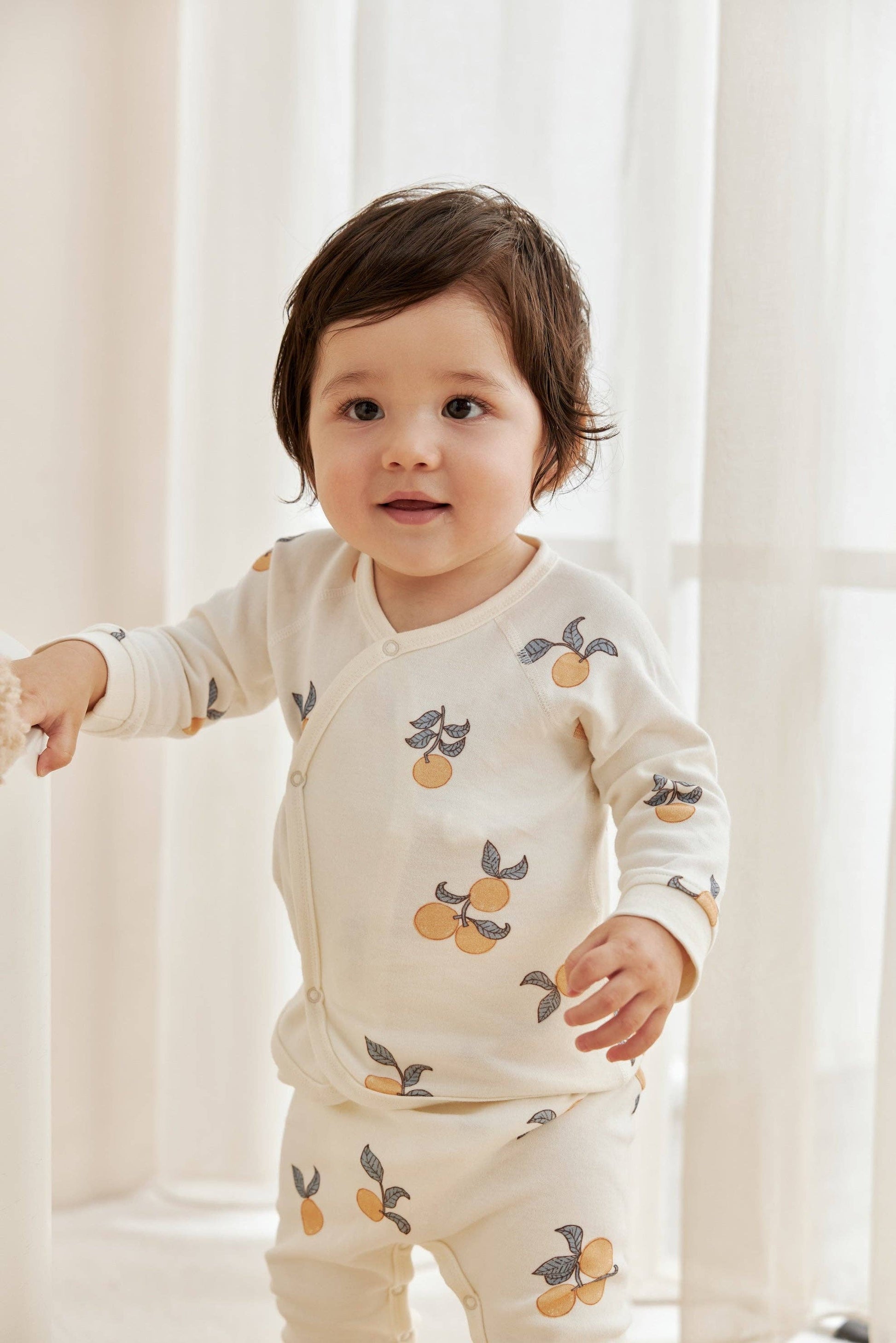 Baby wearing a cream-colored outfit with fruit pattern standing in front of white curtains.