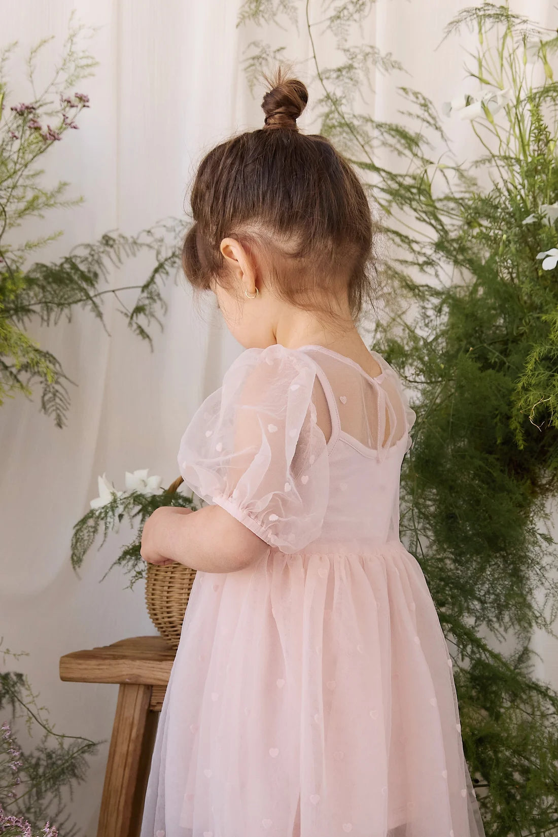 A toddler girl seen from behind wearing a light pink tulle dress with sheer puff sleeves and tiny white polka dots, standing in a styled indoor setting with green plants and a wooden stool.