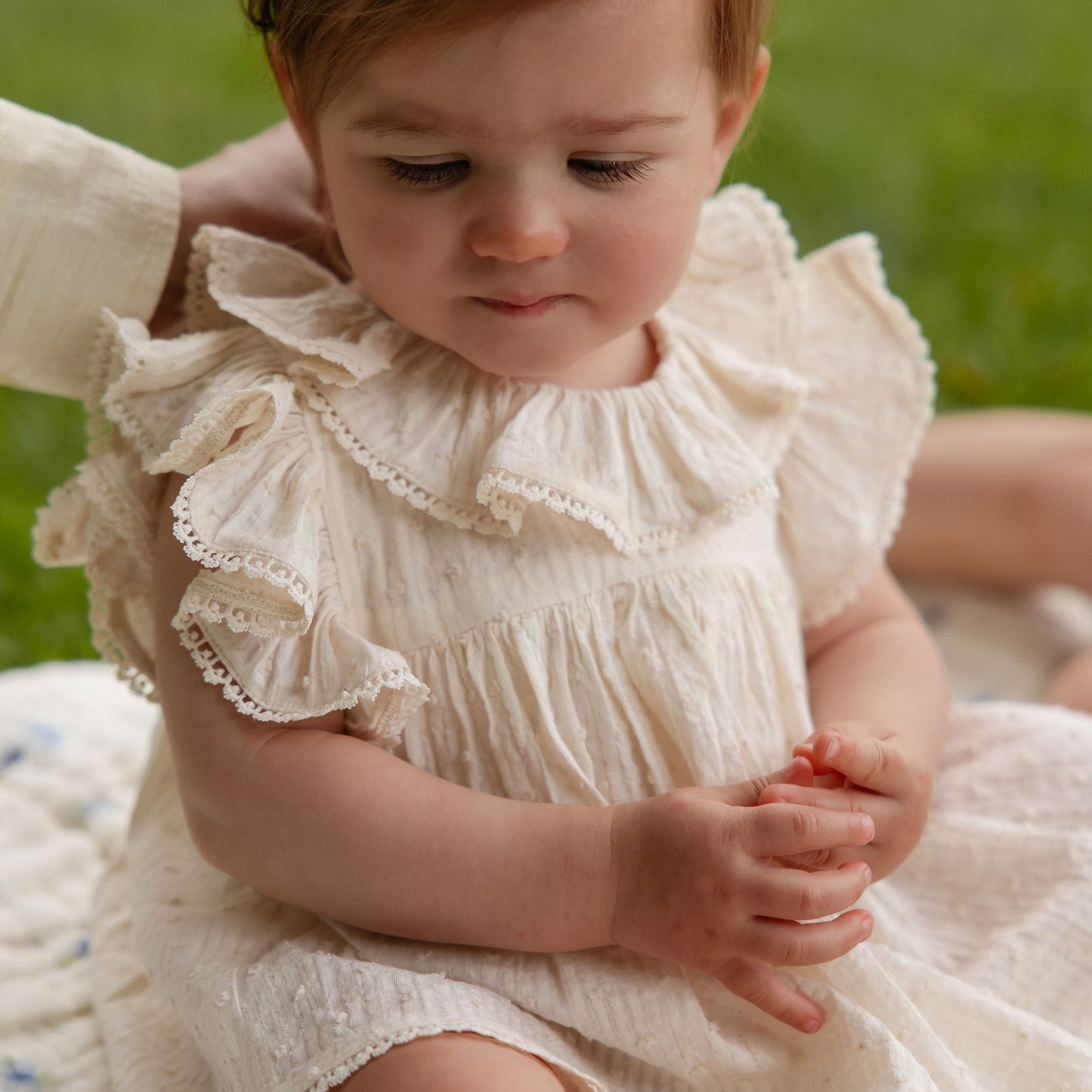 Child wearing a white dress with ruffles sitting on a blanket outdoors.