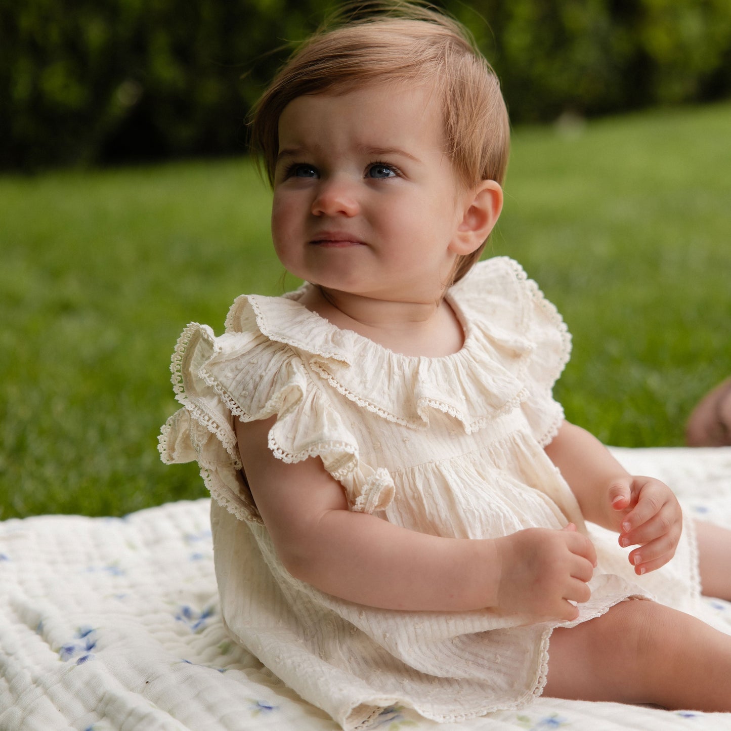 Baby in a ruffled outfit sitting on a blanket in a grassy area