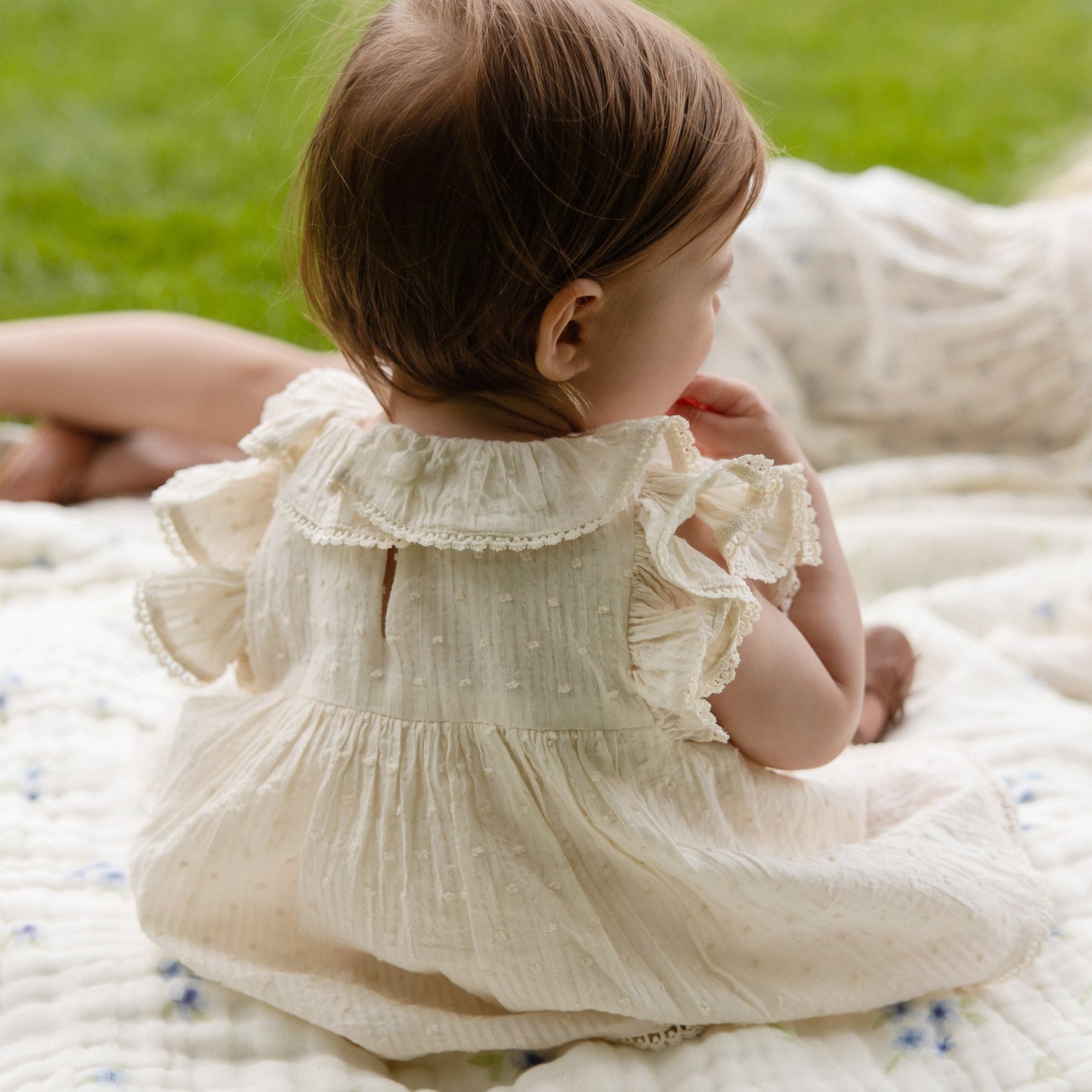 Baby in a white dress sitting on a quilted blanket outdoors.