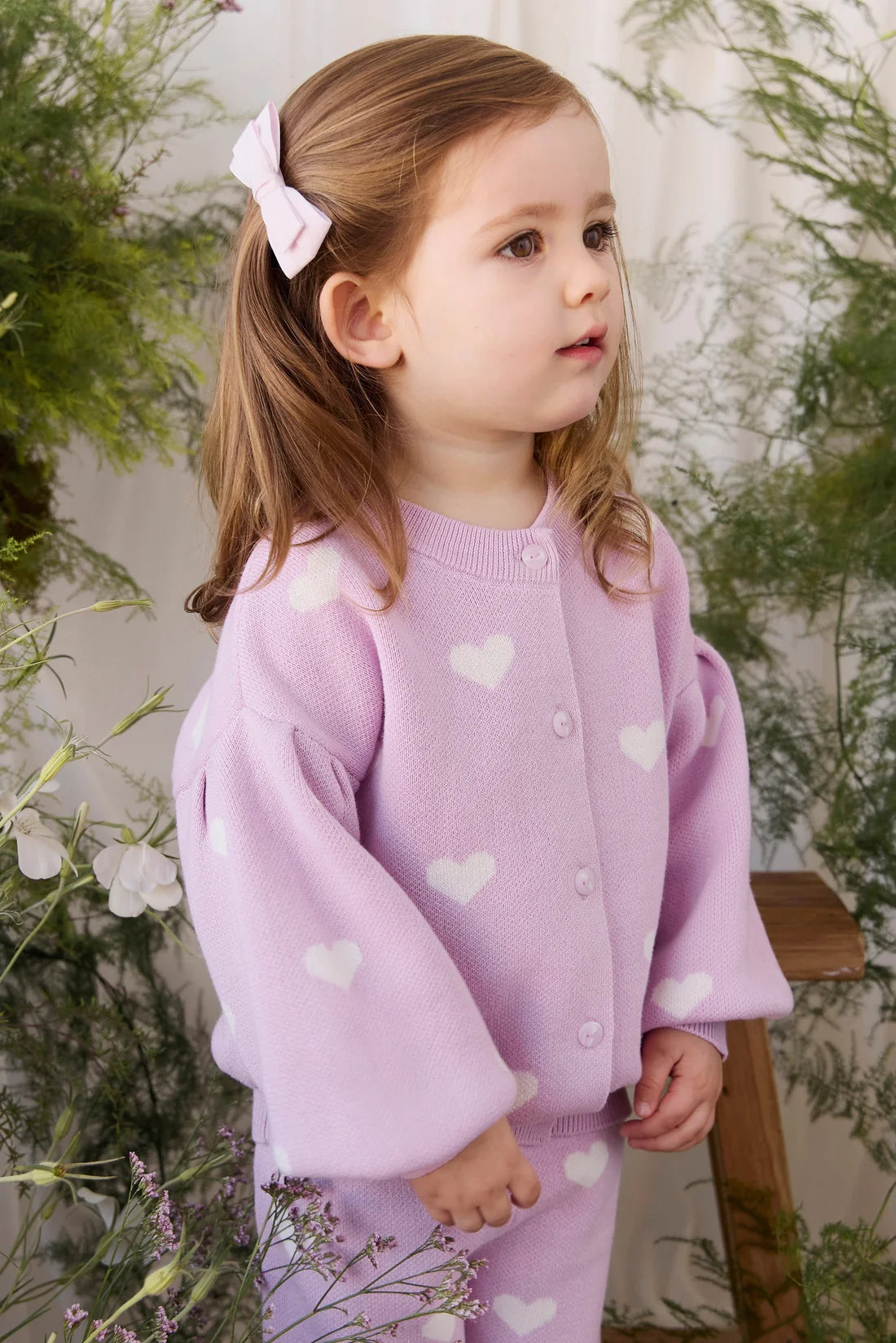 A young child wearing a light purple knit cardigan and matching pants with white heart patterns, standing in front of a soft white curtain and green foliage.