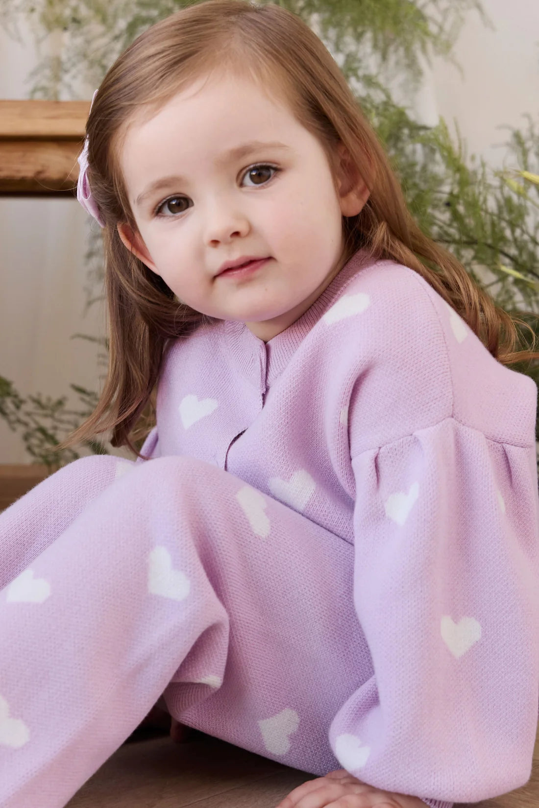 A young child wearing a light purple fleece sleeper or one-piece pajama with white heart patterns, sitting down in a cozy indoor setting.