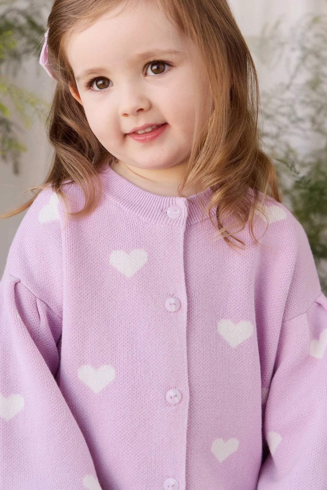 A young girl with light brown hair wearing a lavender knit cardigan sweater with white heart patterns, photographed in a soft-focus indoor setting.
