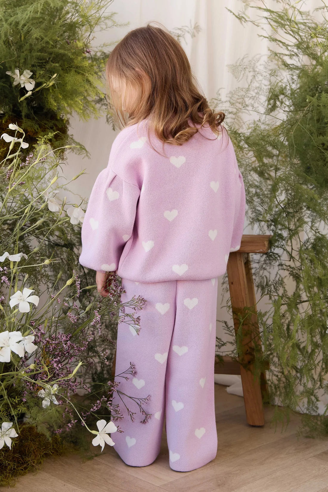A child wearing a light pink ribbed knit two-piece outfit with white heart patterns, photographed from behind in a styled indoor setting with greenery and a wooden stool.