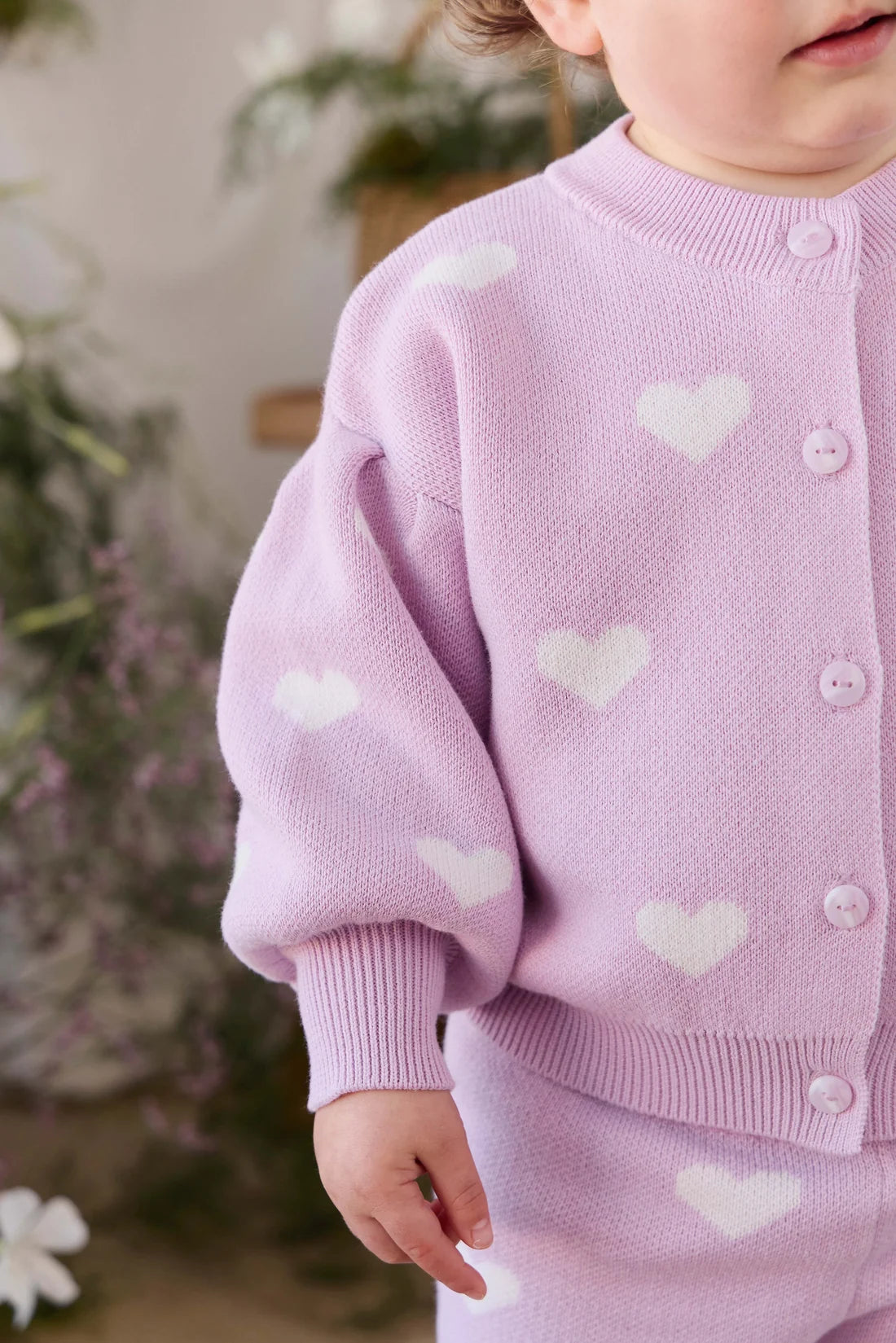 A close-up of a child wearing a light lavender knit cardigan with white heart motifs and matching ribbed cuffs and hem, photographed indoors with a softly blurred floral background.