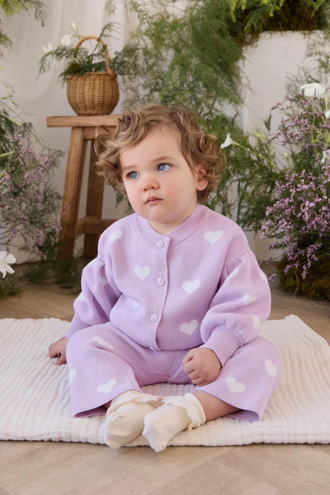 A toddler with curly light brown hair sits on a cream ribbed blanket in a styled indoor setting with greenery and a wooden stool. The child is wearing a lavender cardigan and matching pants covered in white heart patterns, paired with cream tights.