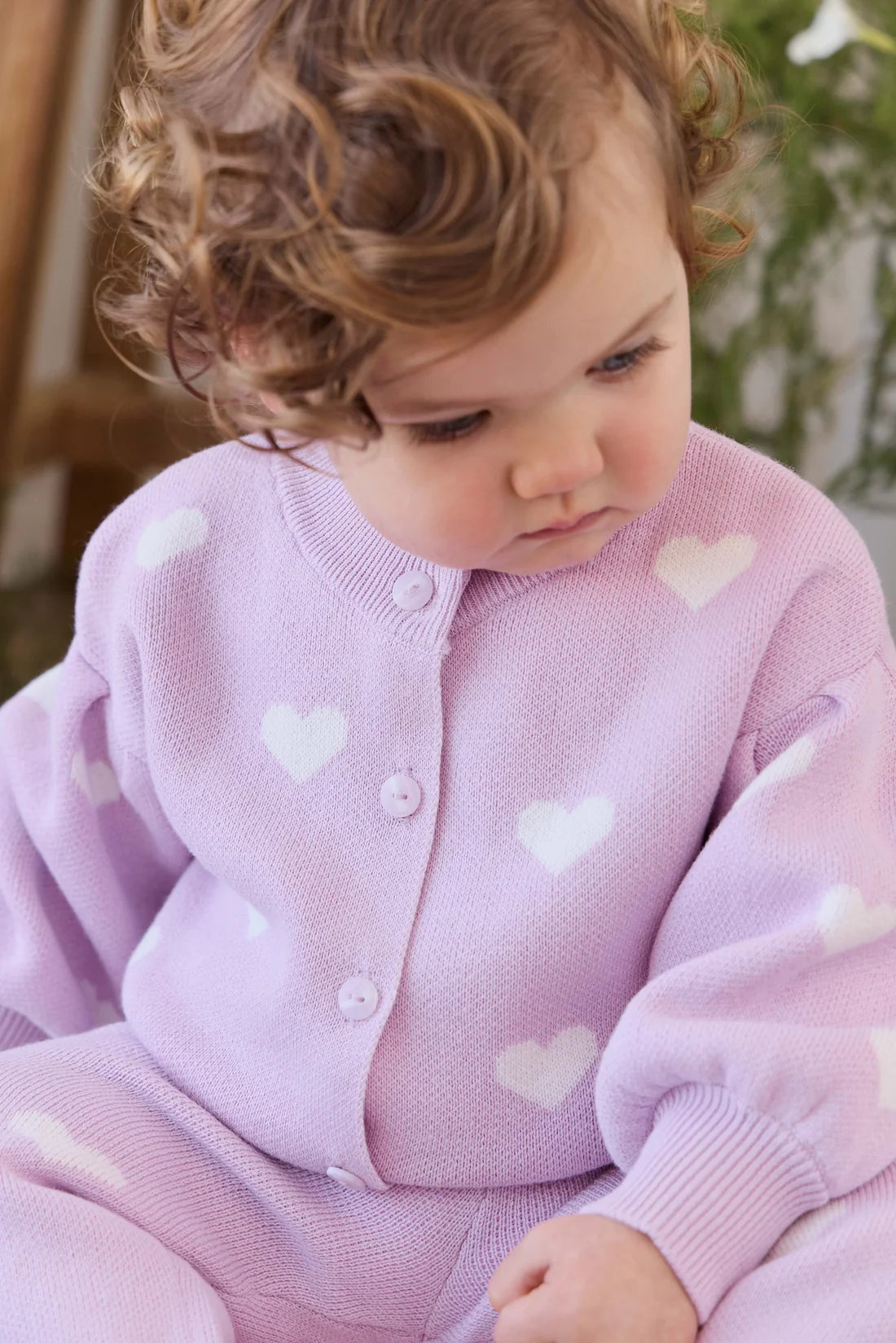 A close-up of a toddler wearing a light purple knit cardigan and matching pants with white heart patterns, looking down thoughtfully.