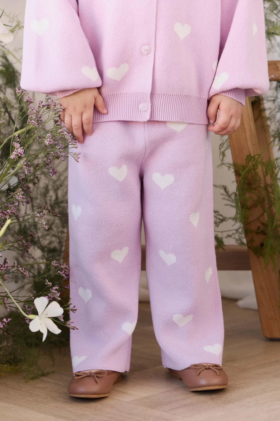 A child wearing a light pink knit cardigan and matching pants with white heart patterns, paired with brown leather shoes, standing in a styled indoor setting with flowers and a wooden chair.