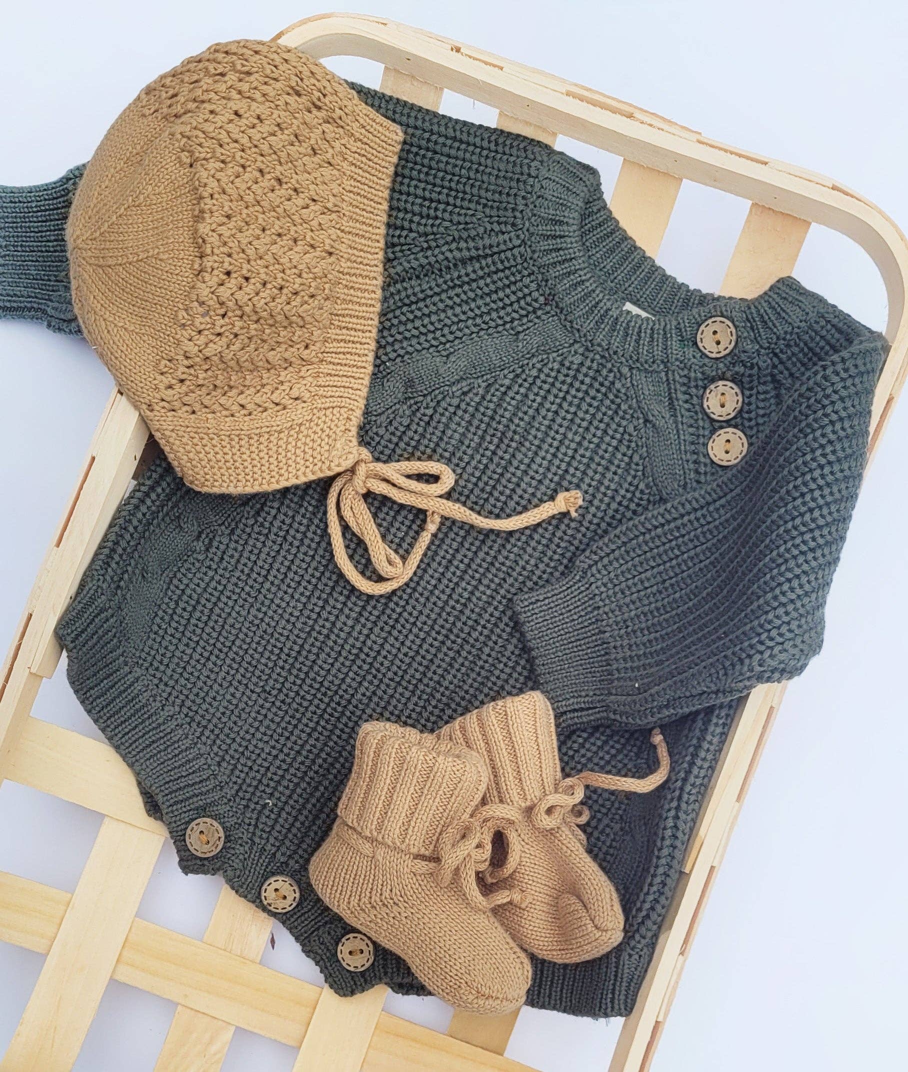 Knitted baby outfit with green sweater, beige hat, and booties on a wooden chair.