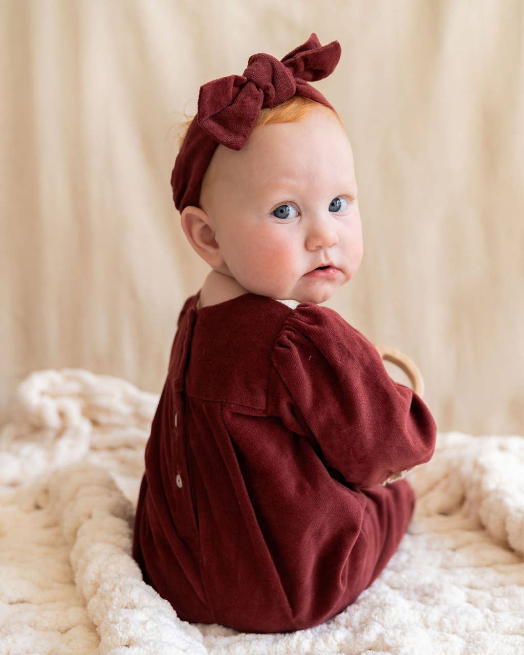 Baby wearing a burgundy outfit and headband sitting on a soft surface.