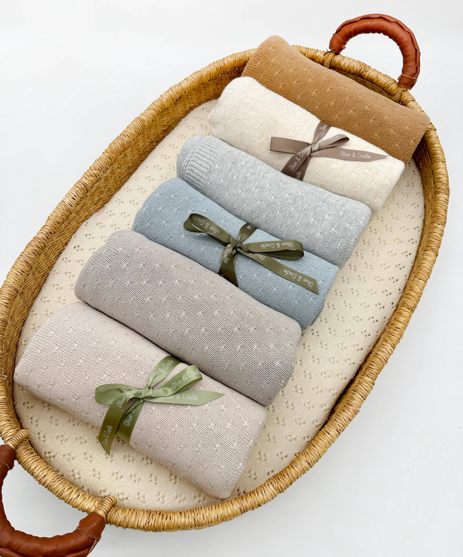 Set of four folded blankets in beige, gray, blue, and brown with green ribbons in a wicker basket on a white background.