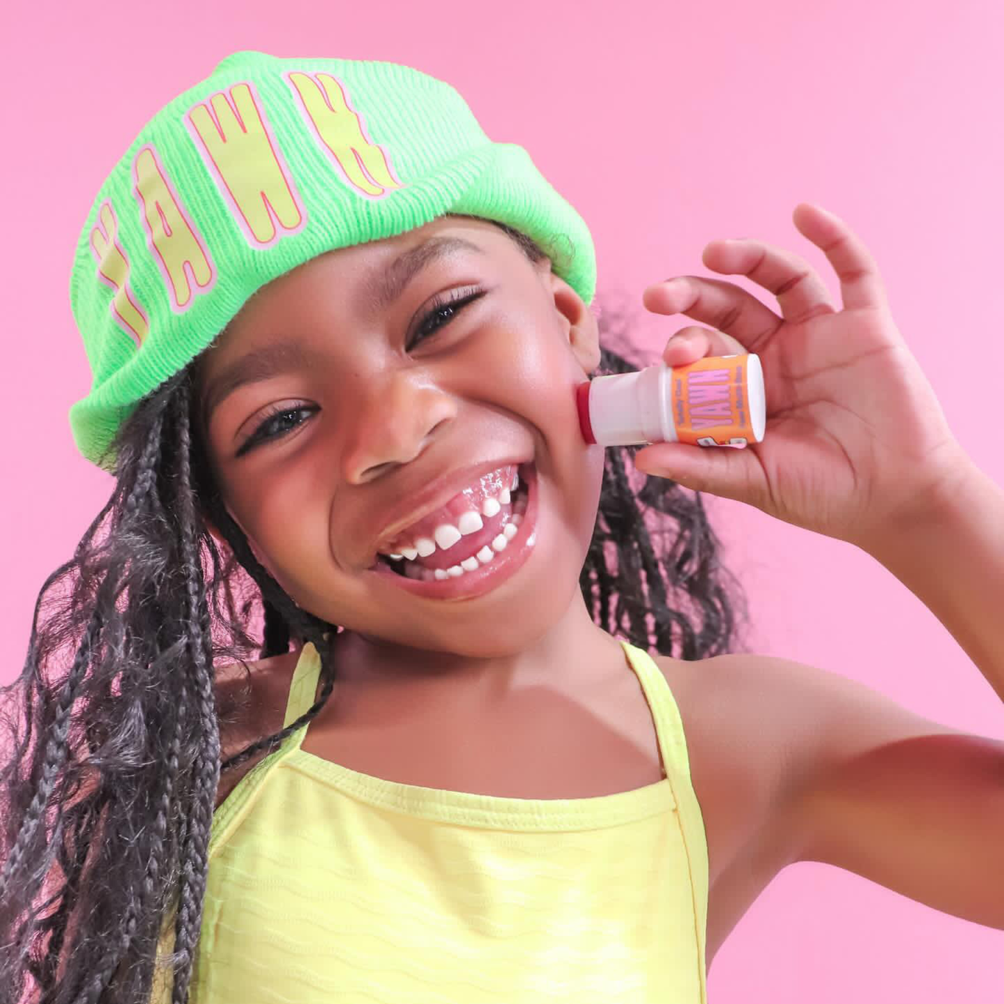 Child wearing a green beanie with 'HAWAII' and applying lip balm against a pink background