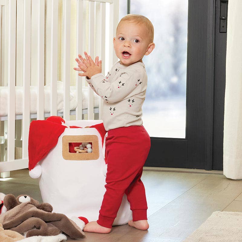 Baby in red pants and white shirt standing next to a potty with a Santa hat