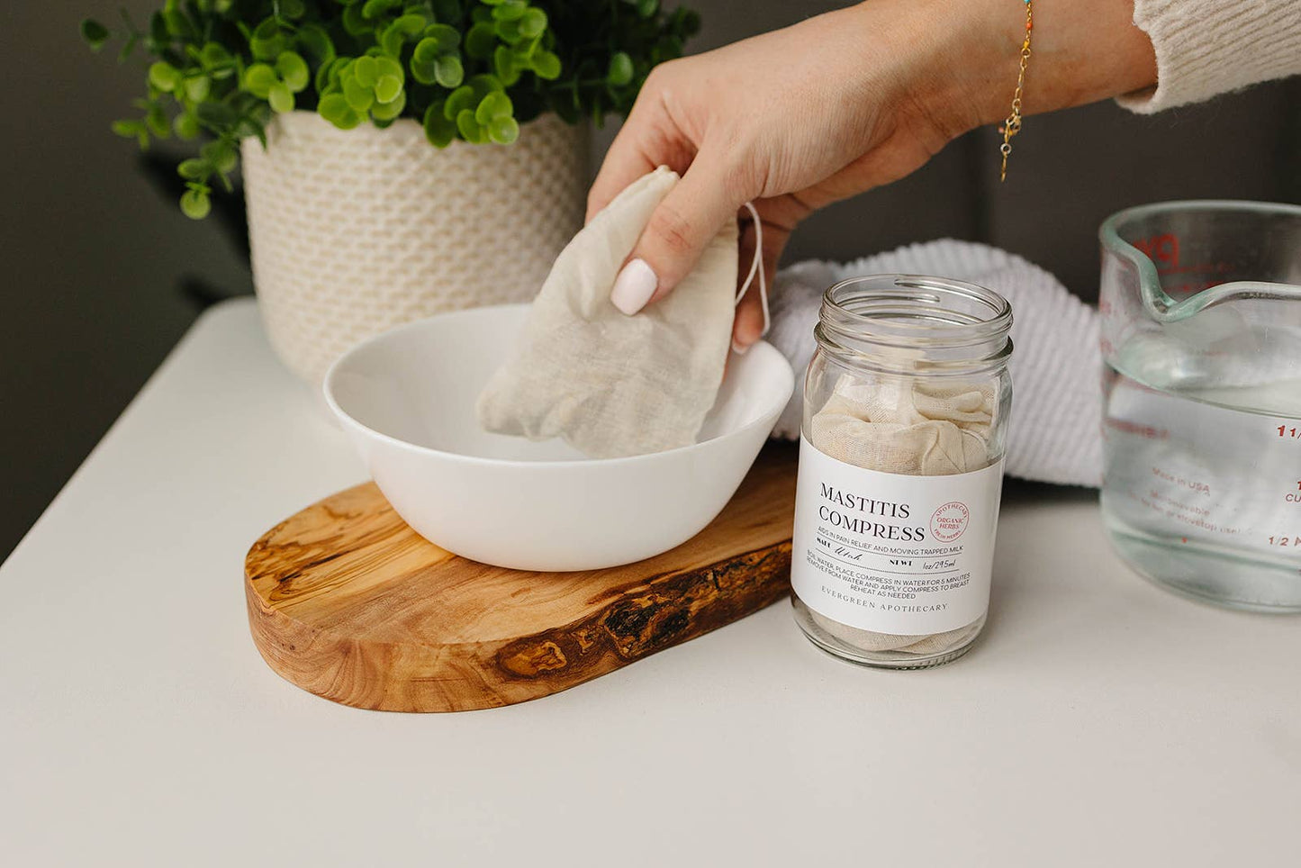 Person holding a compress over a bowl with a jar labeled 'Mastitis Compress' on a table.