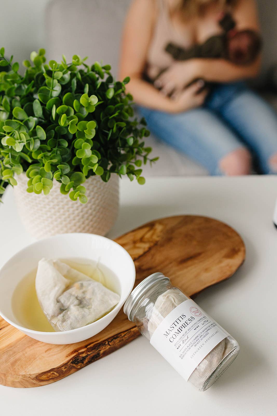 Tea bag in a bowl with a jar of tea leaves on a wooden board, with a blurred background of a person holding a baby.