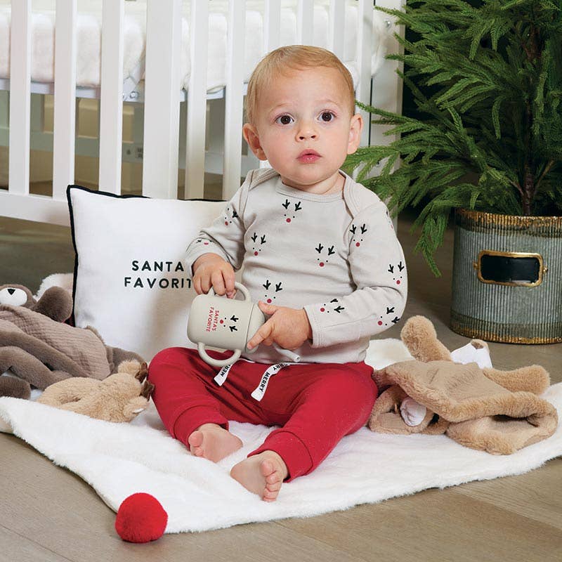Child in red pants and gray shirt holding a mug, surrounded by toys and a 'Santa Favorite' pillow.