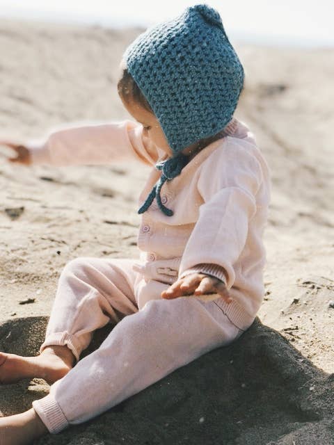 Child wearing a blue knitted bonnet and pink outfit sitting on sand