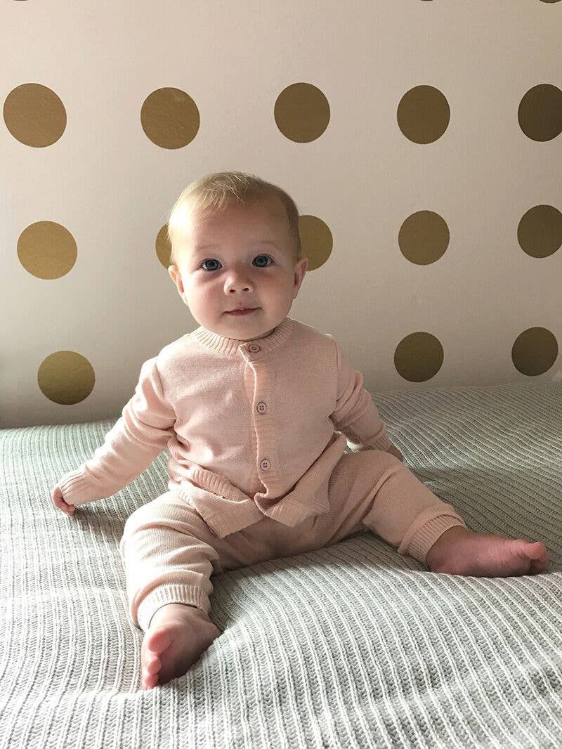 Baby in a pink outfit sitting on a bed with a polka dot wall in the background
