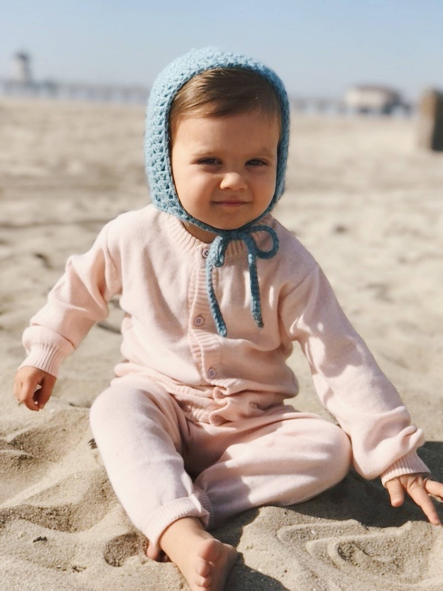 Child wearing a pink outfit and blue bonnet sitting on a sandy beach.