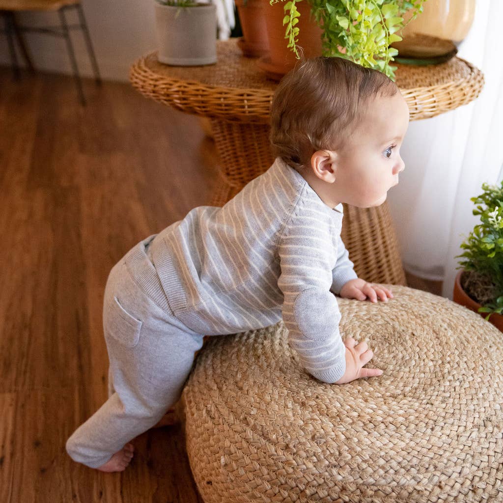 Baby in a striped outfit standing on a woven ottoman in a home setting with plants.
