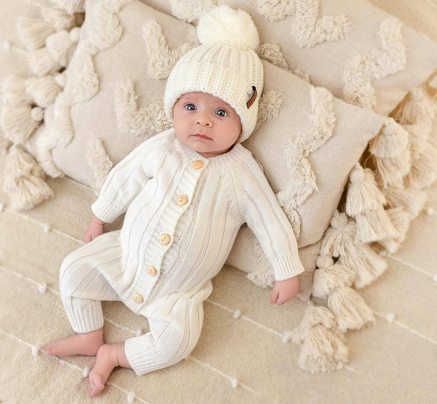 Baby in a white knitted outfit and hat on a soft, textured surface