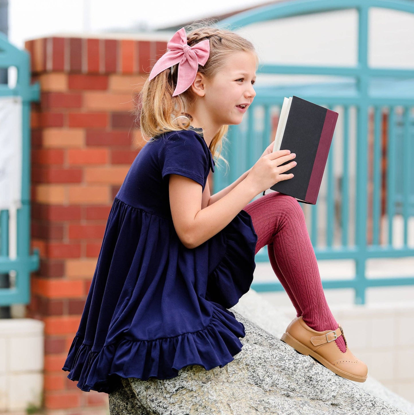 Young girl in a blue dress reading a book outdoors.