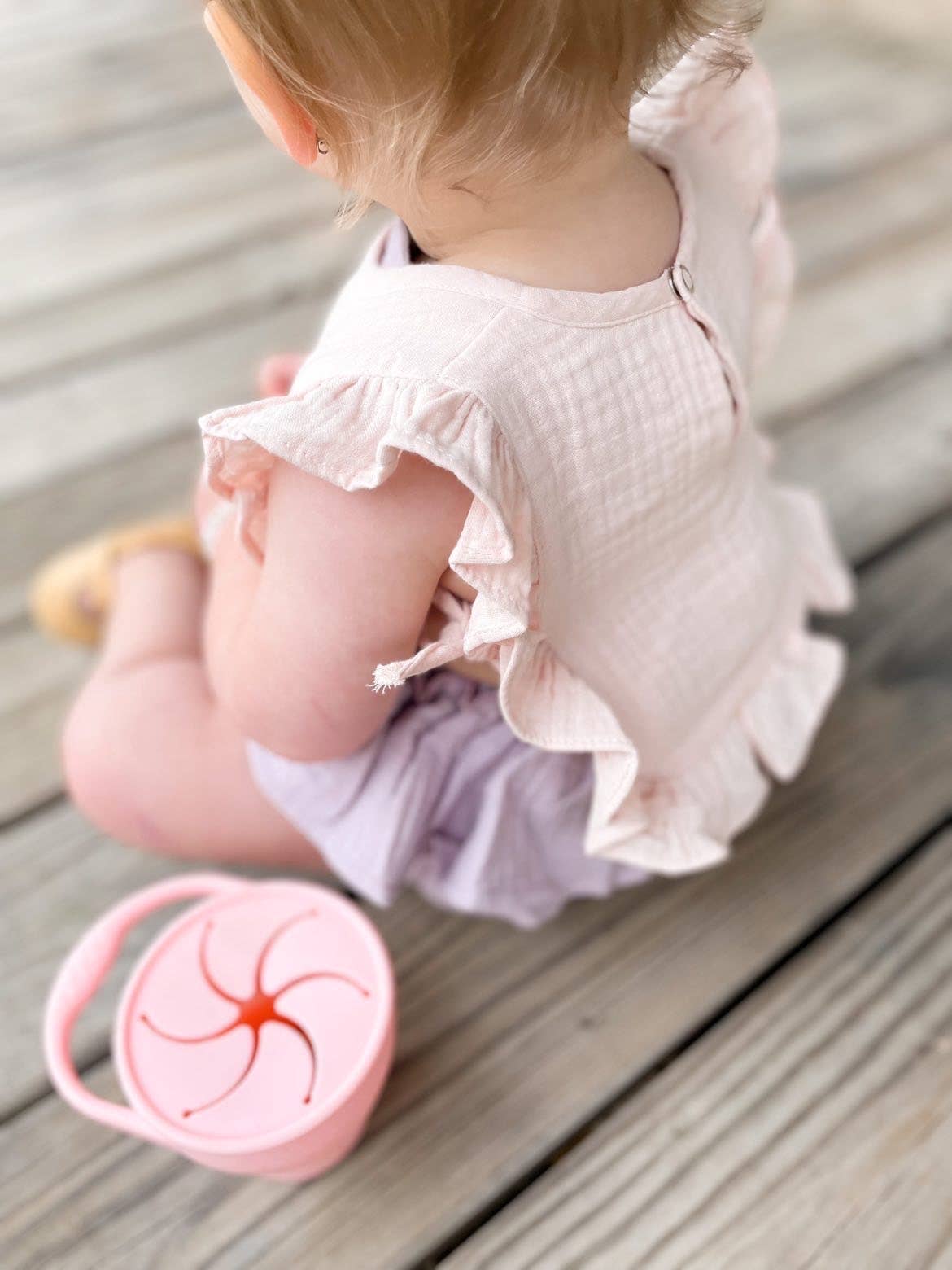 Baby in a light pink outfit sitting on a wooden floor with a pink cup nearby.