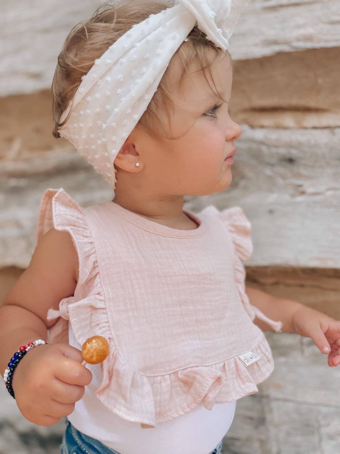 Young child wearing a pink top and white headband, holding a lollipop outdoors.