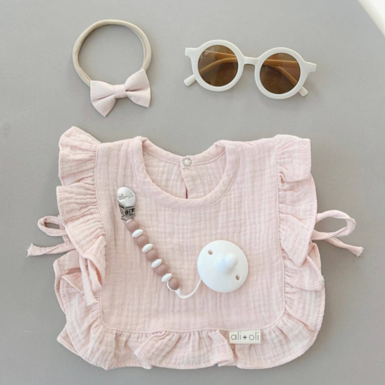 Pink baby bib with accessories including a bow, sunglasses, and teether on a gray background