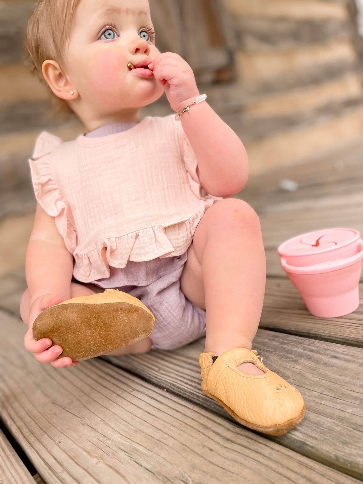 Baby sitting on wooden steps wearing yellow shoes, eating with a pink cup.