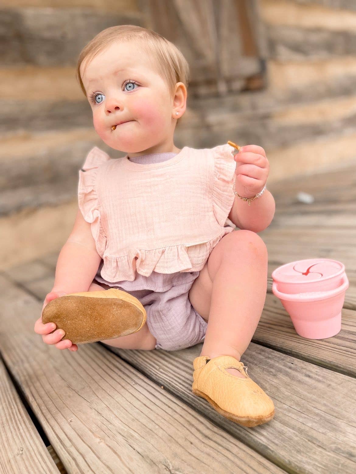 Baby sitting on wooden steps wearing a pink outfit with a pink cup beside her.