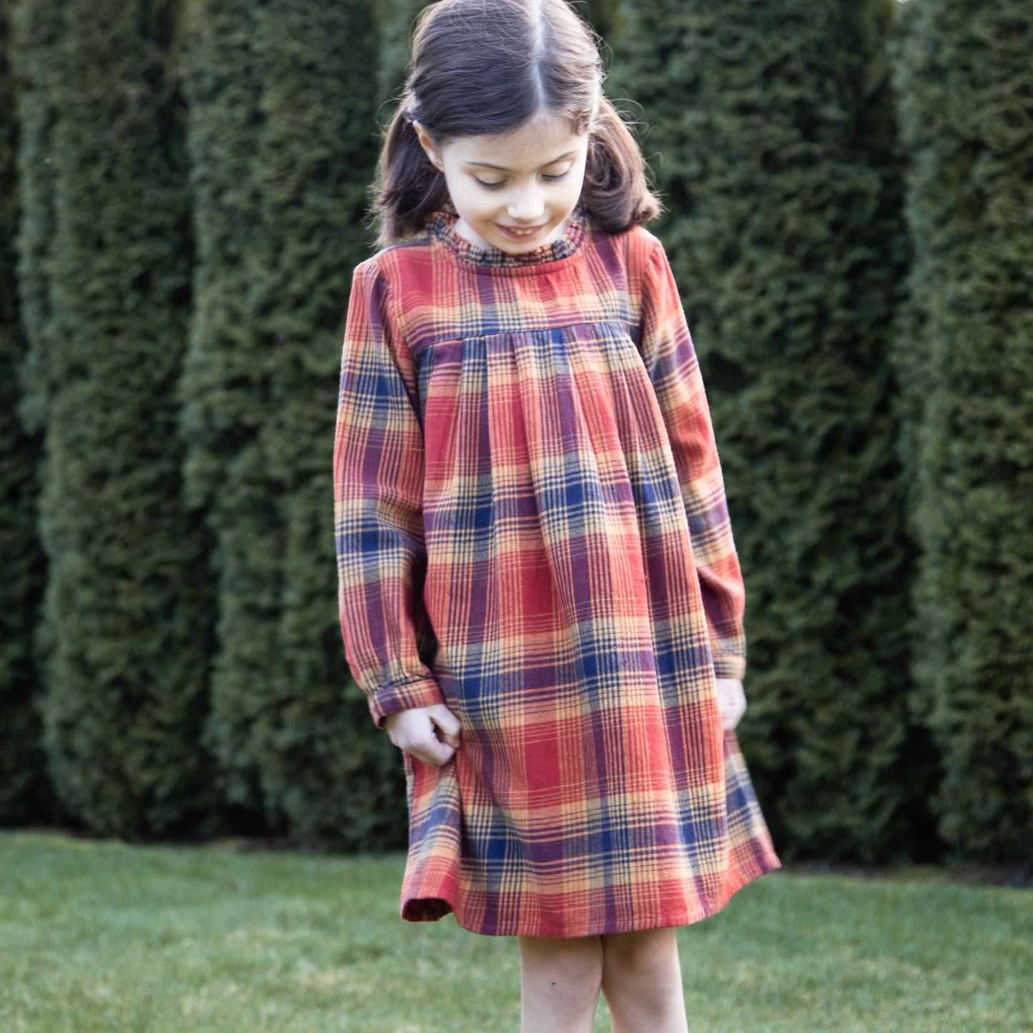 Young girl wearing a plaid dress standing in front of green bushes.
