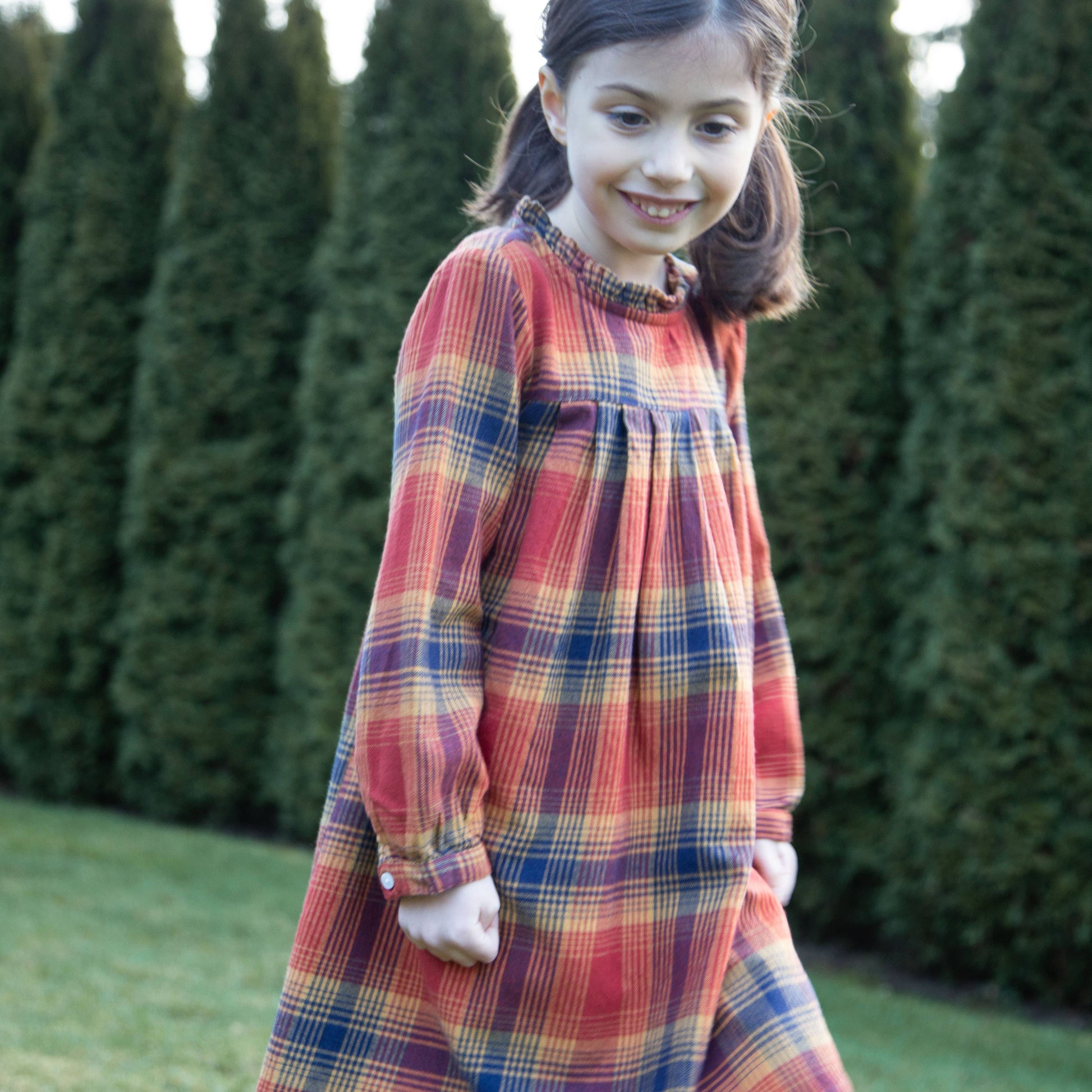 Young girl wearing a plaid dress standing outdoors with greenery in the background