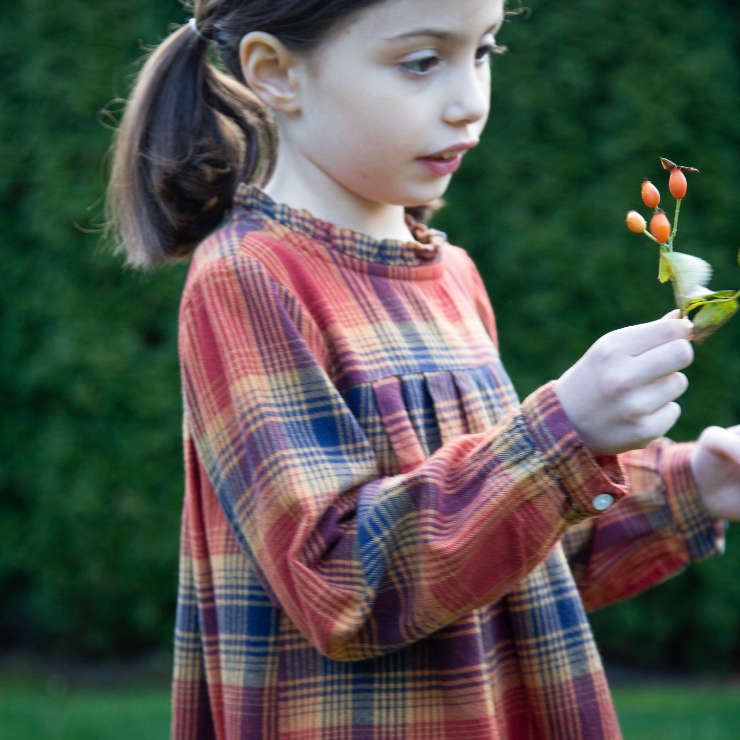 Young girl in a plaid dress holding a small plant with red berries outdoors.