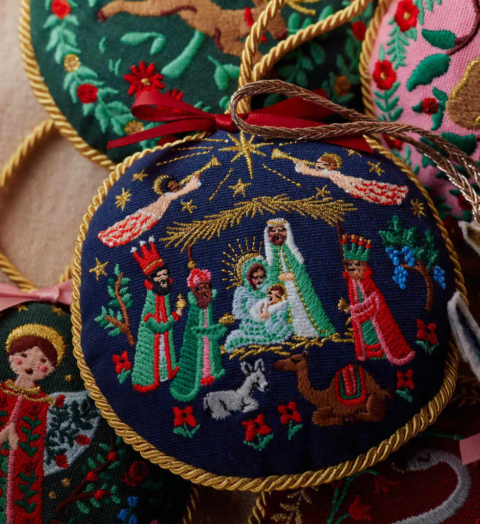Embroidered nativity scene ornament with gold trim on a textured surface.