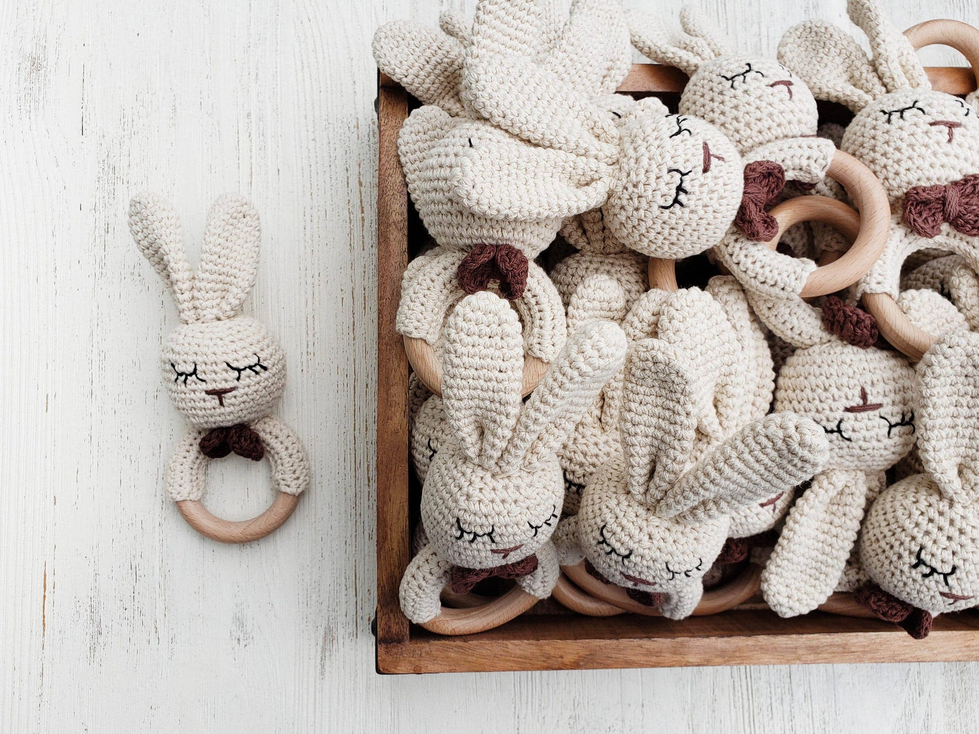 Wooden box filled with crocheted rabbit toys with wooden rings on a light wooden surface.