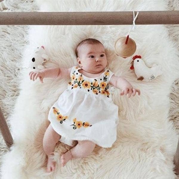 Baby lying on a fluffy white rug wearing a white dress with floral embroidery, surrounded by wooden toys.