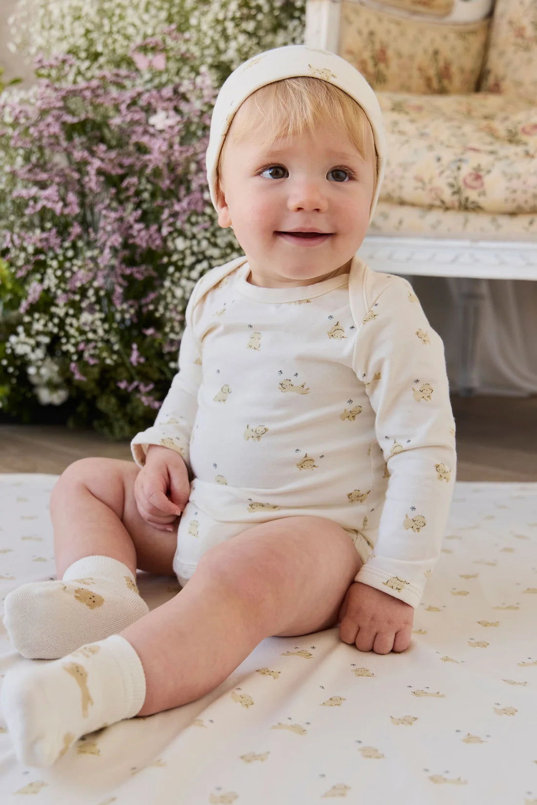 A baby sitting on a cream blanket with a matching printed sheet, wearing a cream long-sleeve bodysuit with small gold teddy bear prints, a matching cream bonnet, and cream socks. The background shows a softly blurred indoor setting with flowers and a chair.