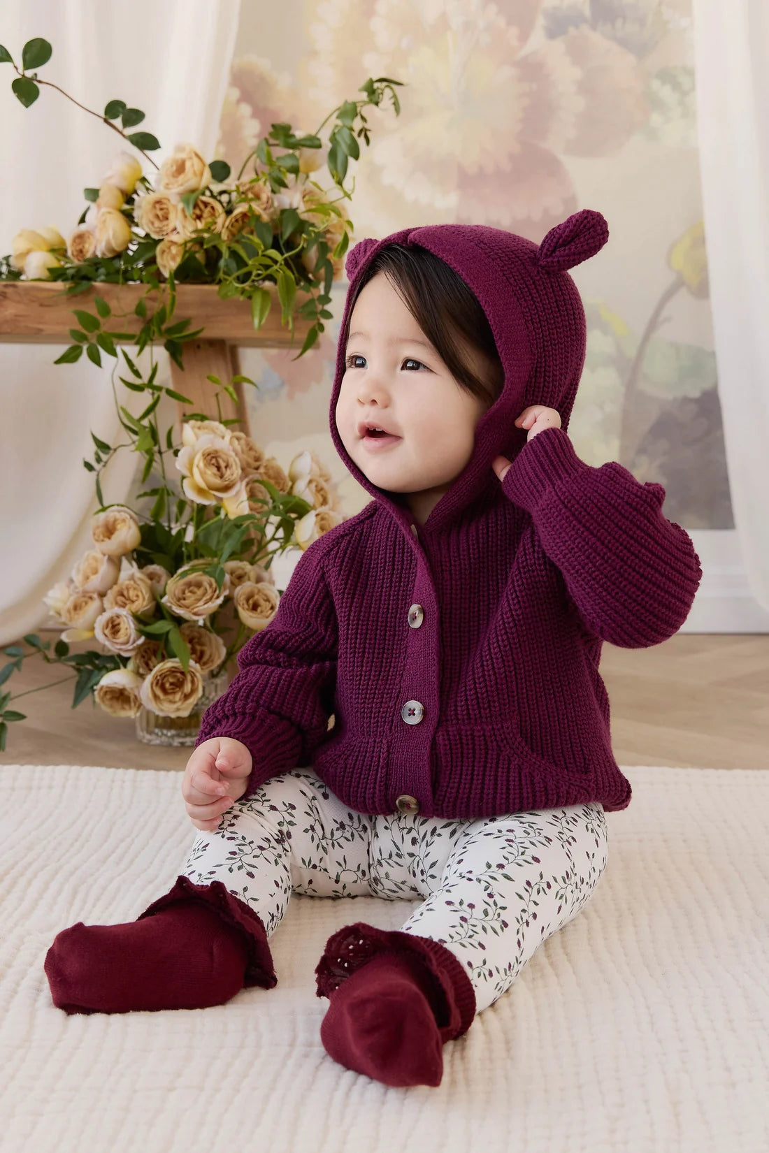 A baby sitting on a cream-colored blanket wearing a deep purple knit cardigan with a matching hood featuring bear ears, patterned white pants, and matching purple booties. The background features a wooden shelf with dried roses and a soft floral print wall.