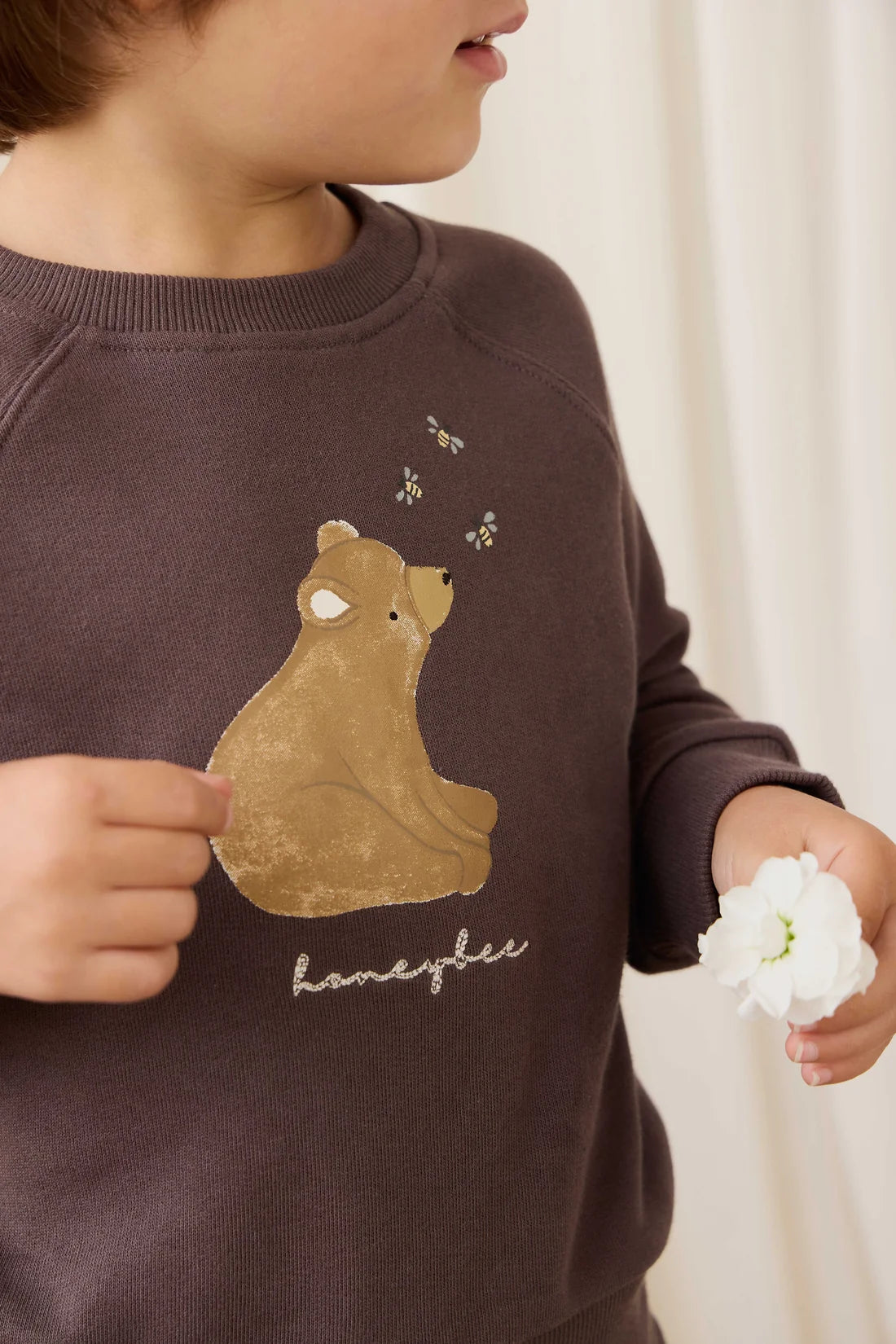 A close-up of a child wearing a dark brown sweatshirt with a graphic of a tan bear, small bees, and the word 'homelee' in white script. The child is holding a white flower against a soft cream curtain background.