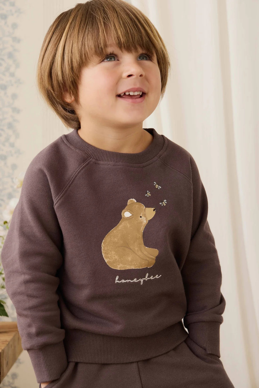 Child wearing a brown sweatshirt with a bear design, standing indoors.