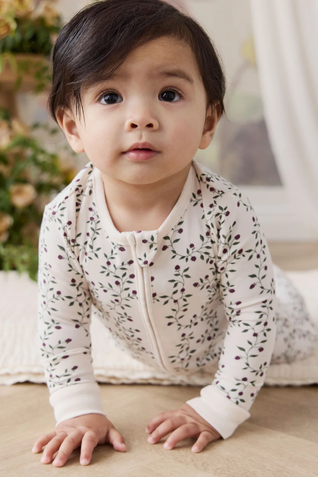 A baby crawling on a wooden floor wearing a cream-colored long-sleeve zip-up sleeper with a delicate green and purple floral pattern.