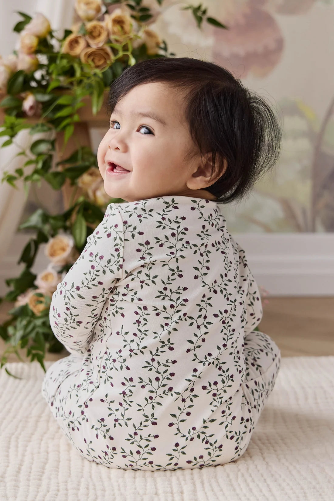 A baby wearing a cream-colored long-sleeve footless sleeper with a green and brown leafy vine pattern, sitting on a textured cream blanket with a floral background.